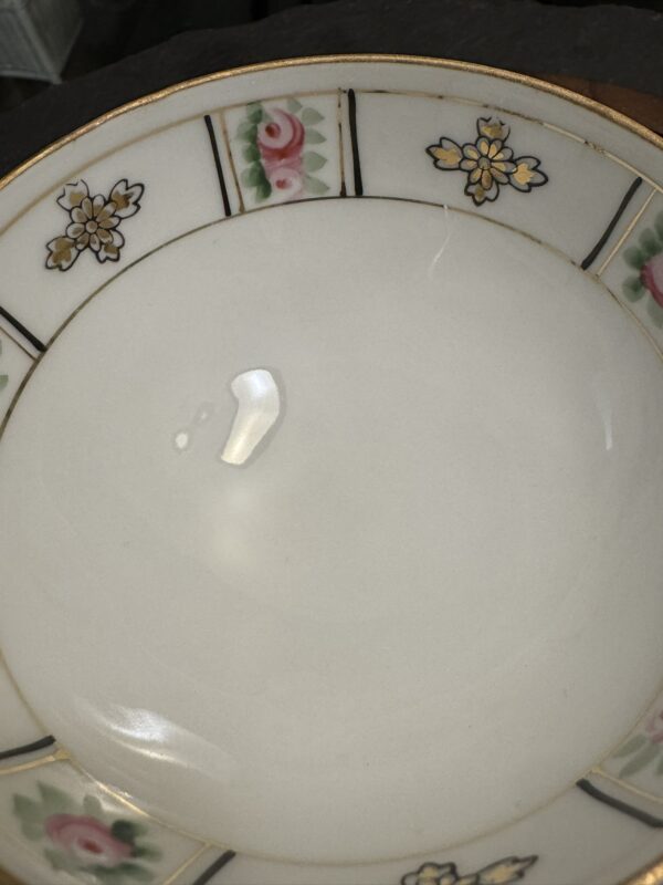 A close-up of a white ceramic plate with a gold rim and floral patterns featuring pink roses and yellow flowers. A small amount of clear liquid, possibly water, is on the plate’s left side. The plate rests on a dark surface.