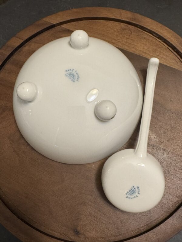 A white ceramic lid with three round rests and a matching ladle sit upside down on a wooden surface. Both show blue maker’s marks with text and a logo. The lid and ladle are smooth and glossy, reflecting light.
