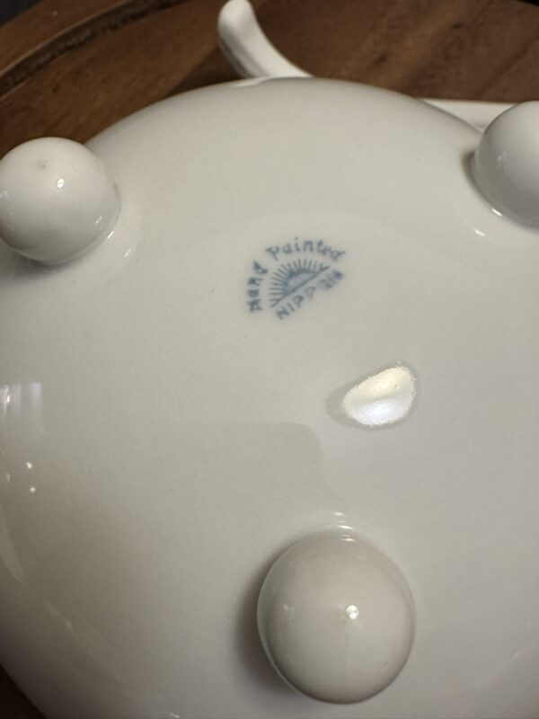 The bottom of a white ceramic object with three small, rounded feet and a handle visible. A blue stamp reads “Hand Painted, Nippon,” with a partial sunburst logo. The surface is glossy and the background appears to be a wooden table.