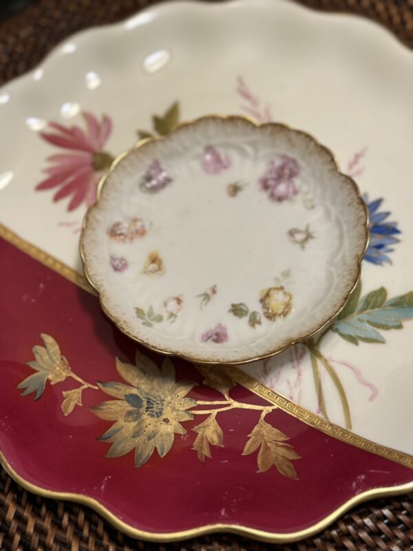 A small, ornate porcelain plate with scalloped gold edges and delicate floral patterns sits atop a larger, decorative plate. The larger plate features vibrant pink, blue, and gold flowers with green leaves on a red and cream background.