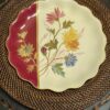 A decorative round plate with a scalloped gold-edged rim sits on a woven rattan placemat. The plate is cream with hand-painted colorful flowers and leaves, and a red section with gold floral accents on the left side.