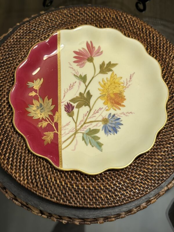 A decorative round plate with a scalloped gold-edged rim sits on a woven rattan placemat. The plate is cream with hand-painted colorful flowers and leaves, and a red section with gold floral accents on the left side.