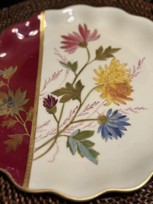 A decorative plate with scalloped edges features colorful flowers—yellow, pink, blue, and purple—on a cream background. The left section is deep red with gold floral accents, divided by a gold border. The plate rests on a woven surface.
