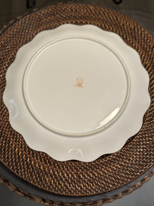 A scalloped-edge white ceramic plate is shown upside down, revealing a faded reddish maker’s mark at the center. The plate rests on a round, woven brown placemat atop a table, with some blurred background elements visible.