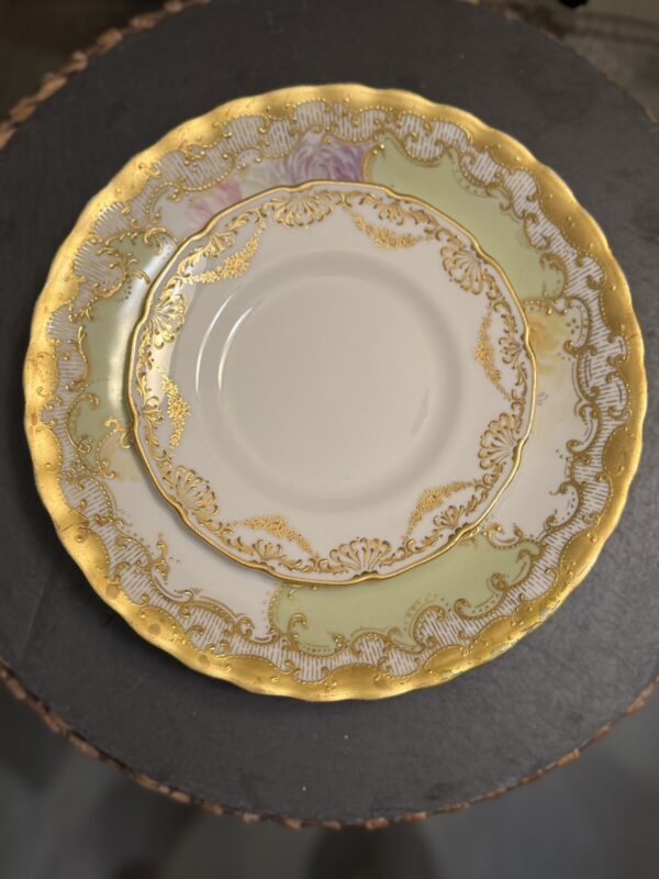 The Sadek Bird Dish features a scalloped gold rim, pale green border, intricate gold patterns, and gold scrollwork encircling a white center, displayed on a dark textured surface.