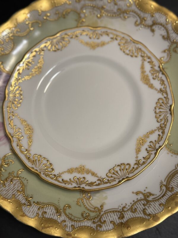 Two ornate porcelain plates overlap. Both have intricate gold patterns and scalloped edges; the smaller plate is a Sadek Bird Dish featuring delicate gold swirls and shell-like accents, while the larger hints at pale green and pink tones.