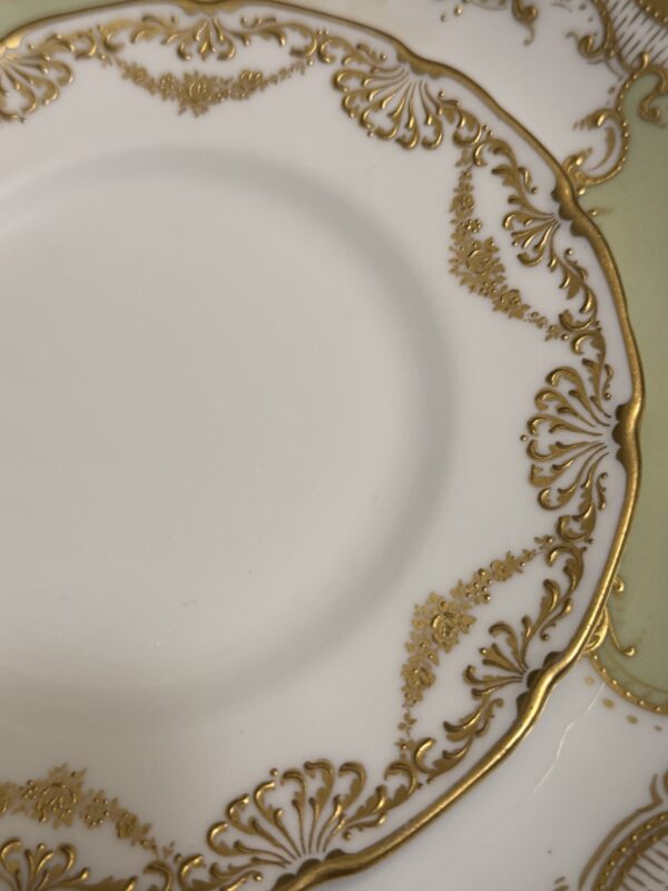 A close-up of the Sadek Bird Dish highlights its ornate gold scrolls, floral motifs, and fan shapes on white porcelain with a pale green border visible in the upper right corner.