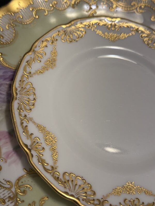 Close-up of ornate Sadek Bird Dish plates stacked together, featuring a white center with intricate gold rims and delicate bird motifs in soft purple and green hues.