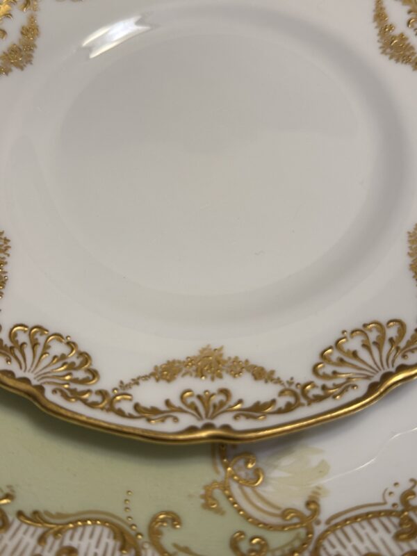 A close-up of two elegant porcelain plates with gold-rimmed details; the top is white with ornate gold, while the bottom resembles a Sadek Bird Dish, featuring a pale green section and sophisticated design.