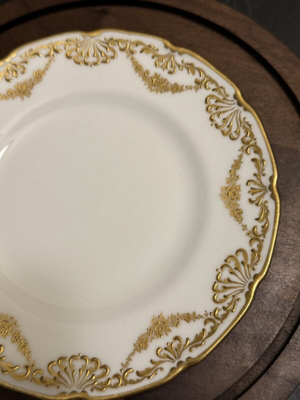 A white porcelain plate with an ornate gold decorative border featuring intricate scrollwork. The plate rests on a wooden charger with a rich dark brown finish. Only part of the plate and charger are visible in the image.