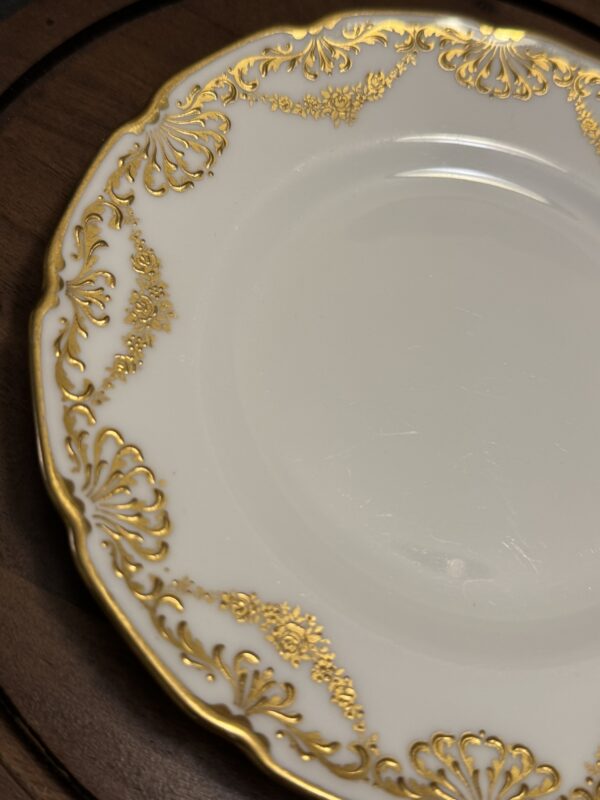 A close-up of a white porcelain plate with an ornate, gold floral and scrollwork border. The plate has a scalloped edge and is set on a dark brown wooden surface. Only part of the plate is visible in the image.