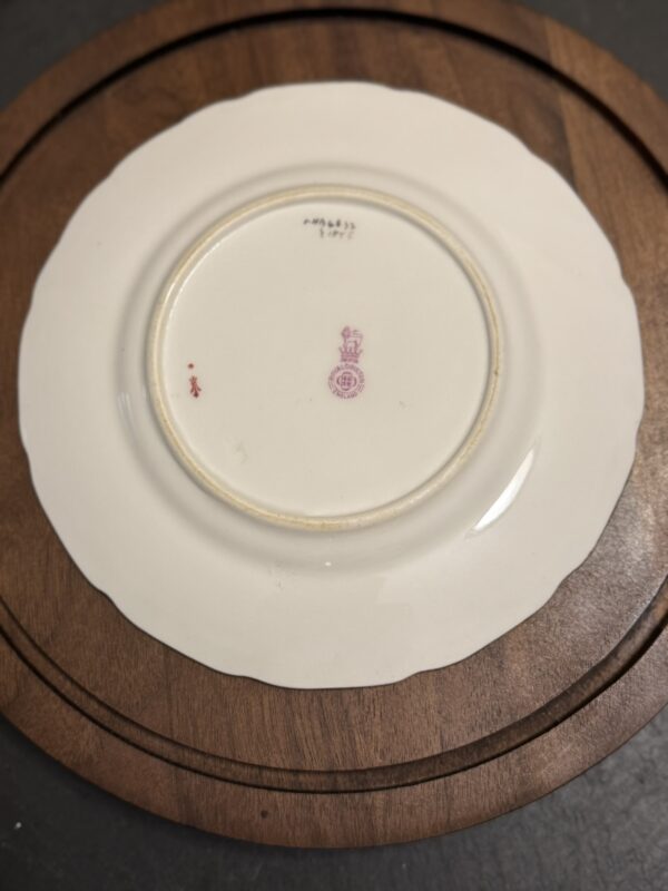 A white ceramic plate lies upside down on a round wooden surface. The plate has scalloped edges and a red maker’s mark with a crown and text at the center of the base, along with other small marks and faded writing near the rim.
