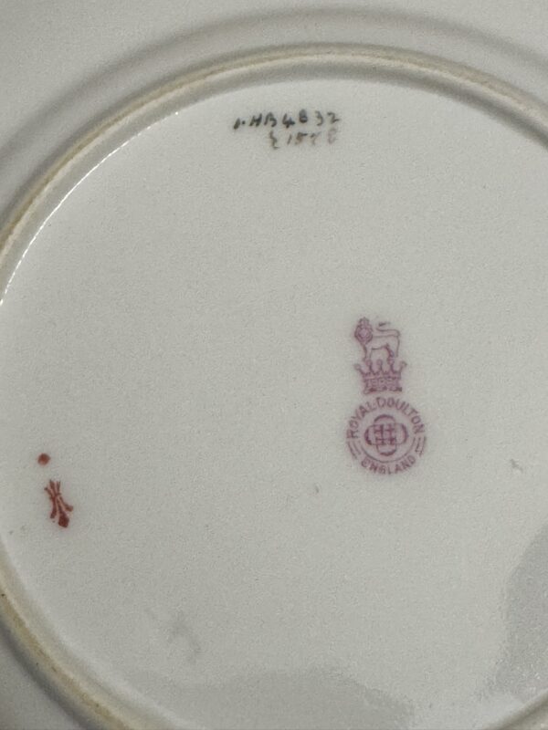 The underside of a white ceramic plate shows a red Royal Doulton England stamp with a lion and crown, a small red symbol, and handwritten numbers and letters in black ink near the rim. The surface appears clean with light wear.