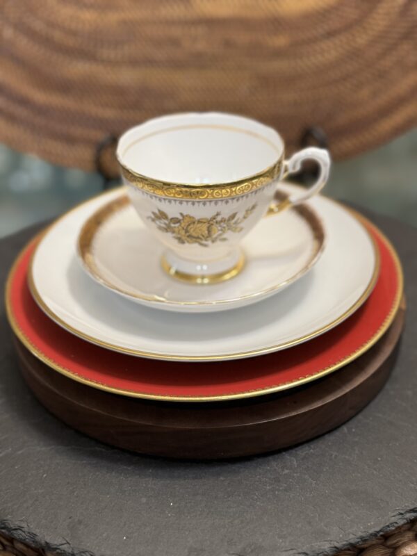 A delicate china teacup with gold floral designs and a matching saucer sit atop two stacked plates—one white with gold trim and one red—arranged on a round, dark slate surface with a woven placemat in the blurred background.