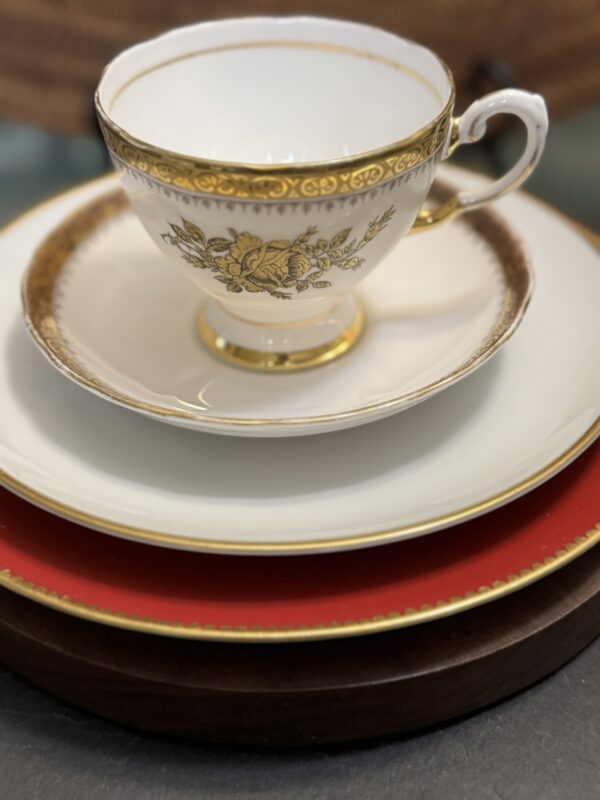 A white porcelain teacup with gold floral detailing sits on a matching saucer atop a cream plate with a gold rim, which rests on a red-rimmed plate. All are stacked on a dark brown charger, creating an elegant, layered table setting.