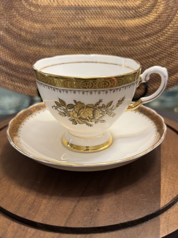A delicate white porcelain teacup with a gold floral design and ornate gold trim sits on a matching saucer. The set rests on a round wooden surface with a woven, textured background behind it.