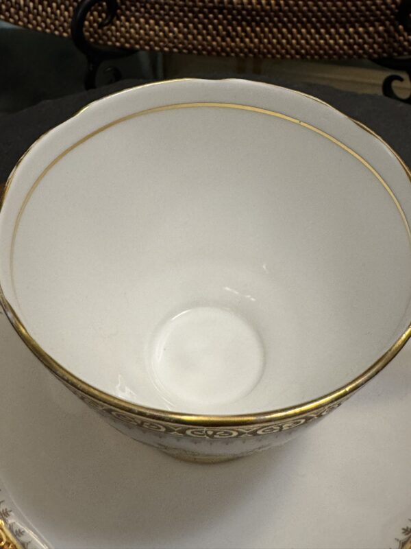 A close-up photo of an empty white porcelain teacup with a thin gold rim, placed on a matching saucer. The teacup features a decorative gold pattern near the rim. The background includes a dark cloth and a woven texture.