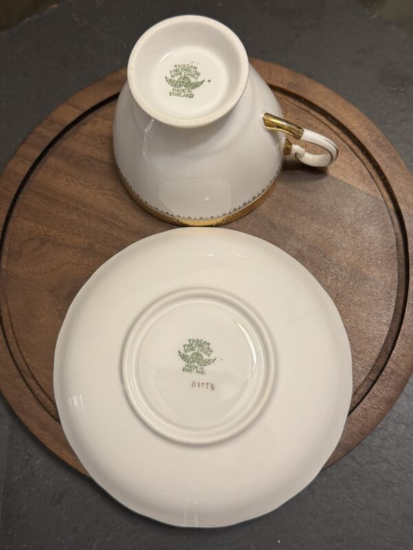 A white teacup and saucer are displayed upside down on a round wooden board. Both have green “Tuscan Fine English Bone China” stamps and gold details along the edges and handle. The saucer also has a red alphanumeric code beneath the stamp.