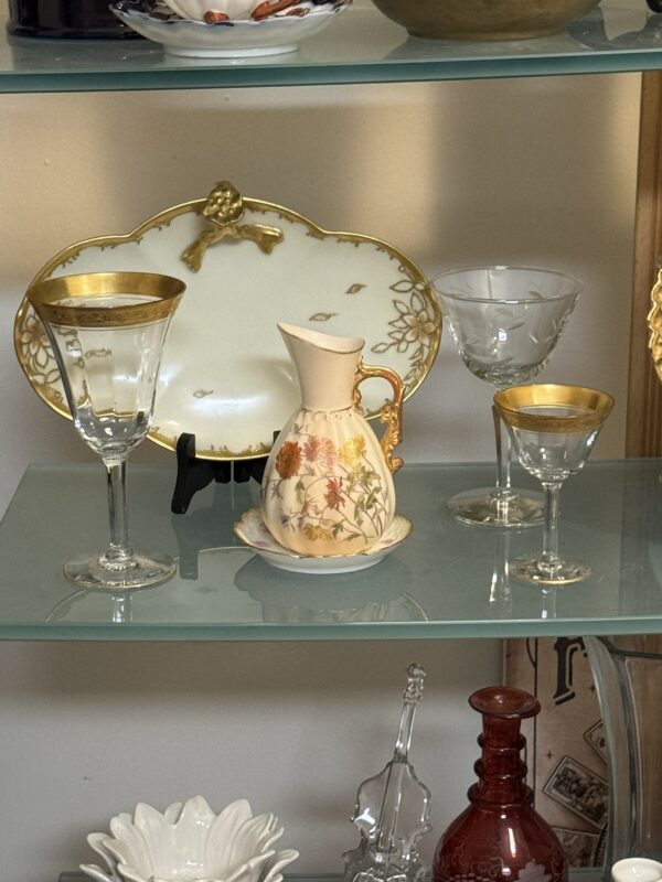 A glass shelf displays a decorative pitcher with floral designs, two wine glasses, one cocktail glass, and a white porcelain dish with gold trim and a raised handle shaped like a flower or cherub. More glassware and ceramics are visible on shelves below.