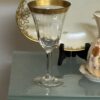 A clear wine glass with a wide gold rim is displayed on a glass shelf. Behind the glass is a decorative plate with gold detailing and floral designs, and to the right, part of a vase with orange and green floral patterns is visible.