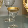 A small, elegant glass with a gold rim sits on a glass shelf. Behind it, there is a larger, clear etched glass with a decorative pattern. Gold-trimmed plates are partially visible at the sides, framing the glasses within a display cabinet.