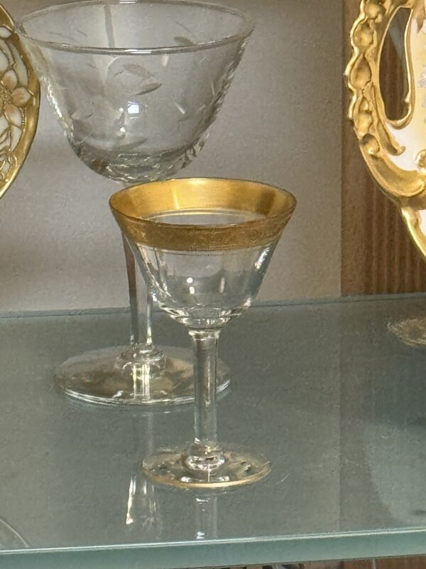 A small, elegant glass with a gold rim sits on a glass shelf. Behind it, there is a larger, clear etched glass with a decorative pattern. Gold-trimmed plates are partially visible at the sides, framing the glasses within a display cabinet.