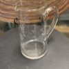 A clear glass pitcher with a handle and spout sits on a dark, round surface. The pitcher is etched with a pattern of grapes and leaves. In the background, there is a woven, circular placemat. The setting appears well-lit and indoors.