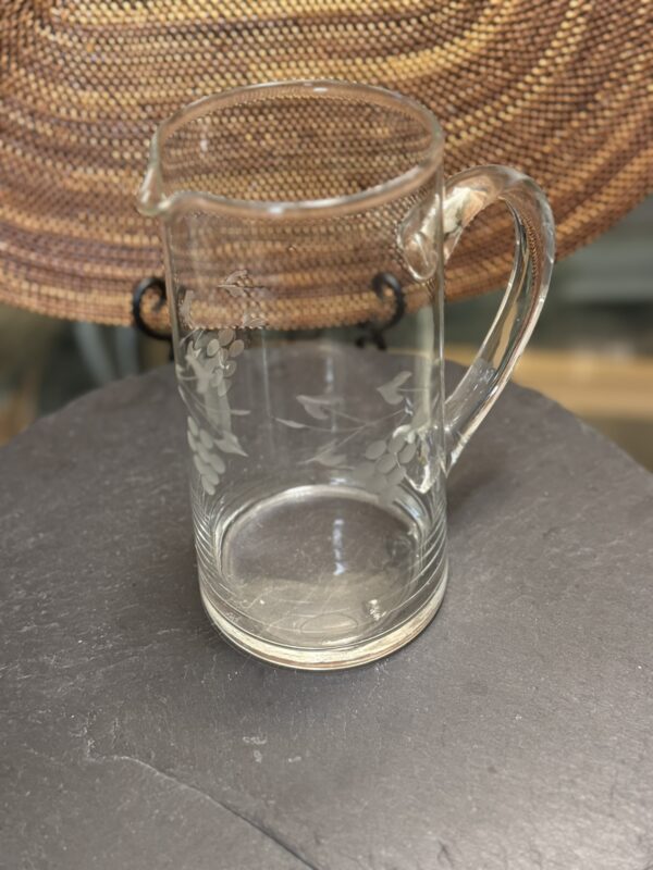 A clear glass pitcher with a handle and spout sits on a dark, round surface. The pitcher is etched with a pattern of grapes and leaves. In the background, there is a woven, circular placemat. The setting appears well-lit and indoors.