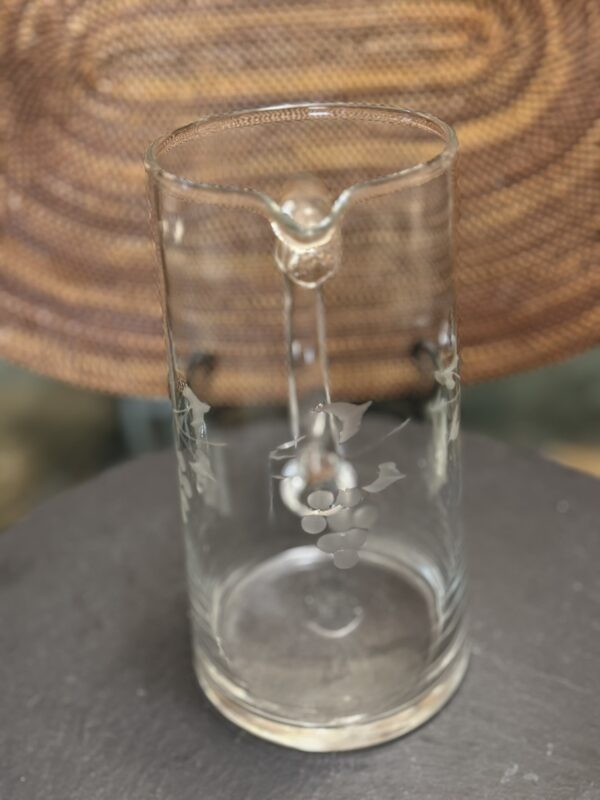 A clear glass pitcher with an etched grapevine design, featuring a bunch of grapes and leaves. The pitcher has a pour spout and is placed on a dark surface with a woven, circular-patterned background.