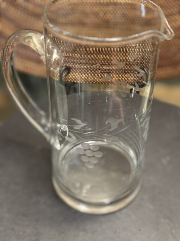 A clear glass pitcher with an etched grapevine design, featuring grape clusters and leaves. The pitcher has a sturdy handle and a spout. It is placed on a dark surface, with a woven background visible behind it.
