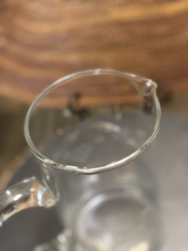 A clear glass pitcher with a chipped rim and a pouring spout sits on a gray surface. The background is blurred, showing a woven, brown material. The damage on the rim is visible along the top edge of the pitcher.