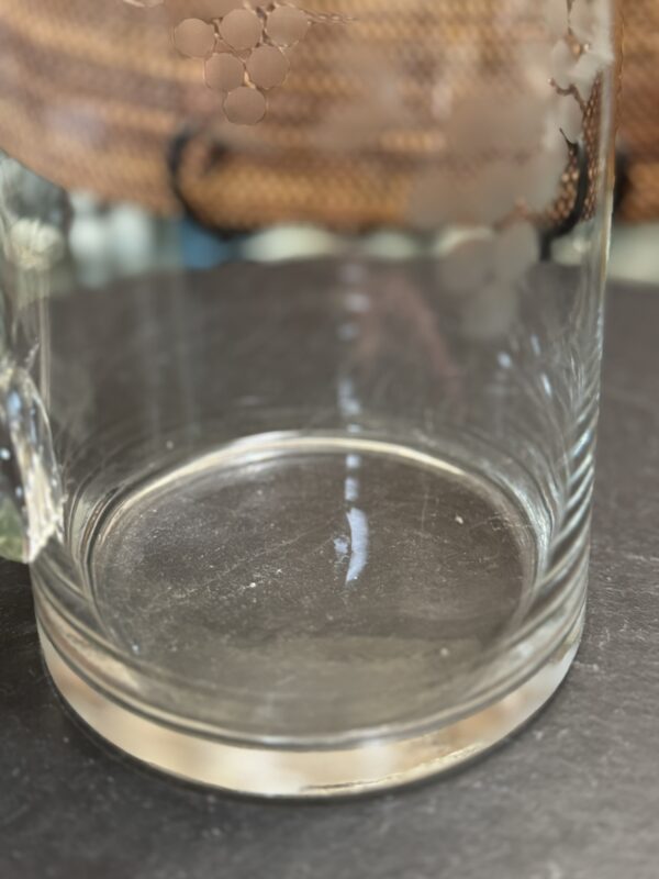 A close-up of the bottom part of a clear glass mug with a handle, placed on a dark surface. The glass has a faint, etched grapevine pattern. The background is blurred, showing a warm, textured surface, possibly wicker or rattan.