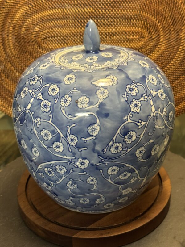 A round, blue porcelain jar with a small lid sits on a wooden base. The jar is decorated with intricate white floral patterns and delicate vines, creating an elegant design. A woven wicker background is visible behind the jar.