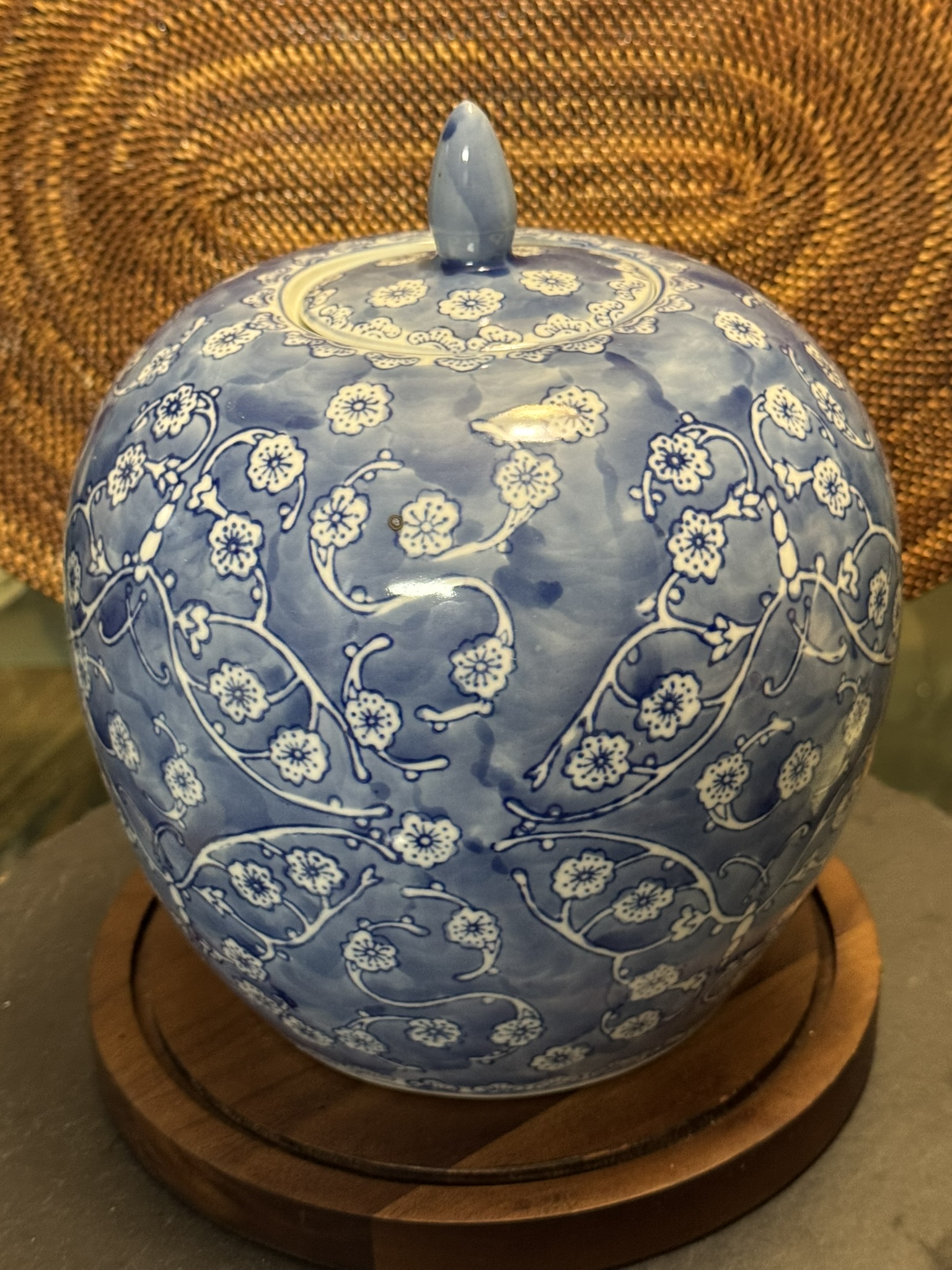A round, blue porcelain jar with a small lid sits on a wooden base. The jar is decorated with intricate white floral patterns and delicate vines, creating an elegant design. A woven wicker background is visible behind the jar.