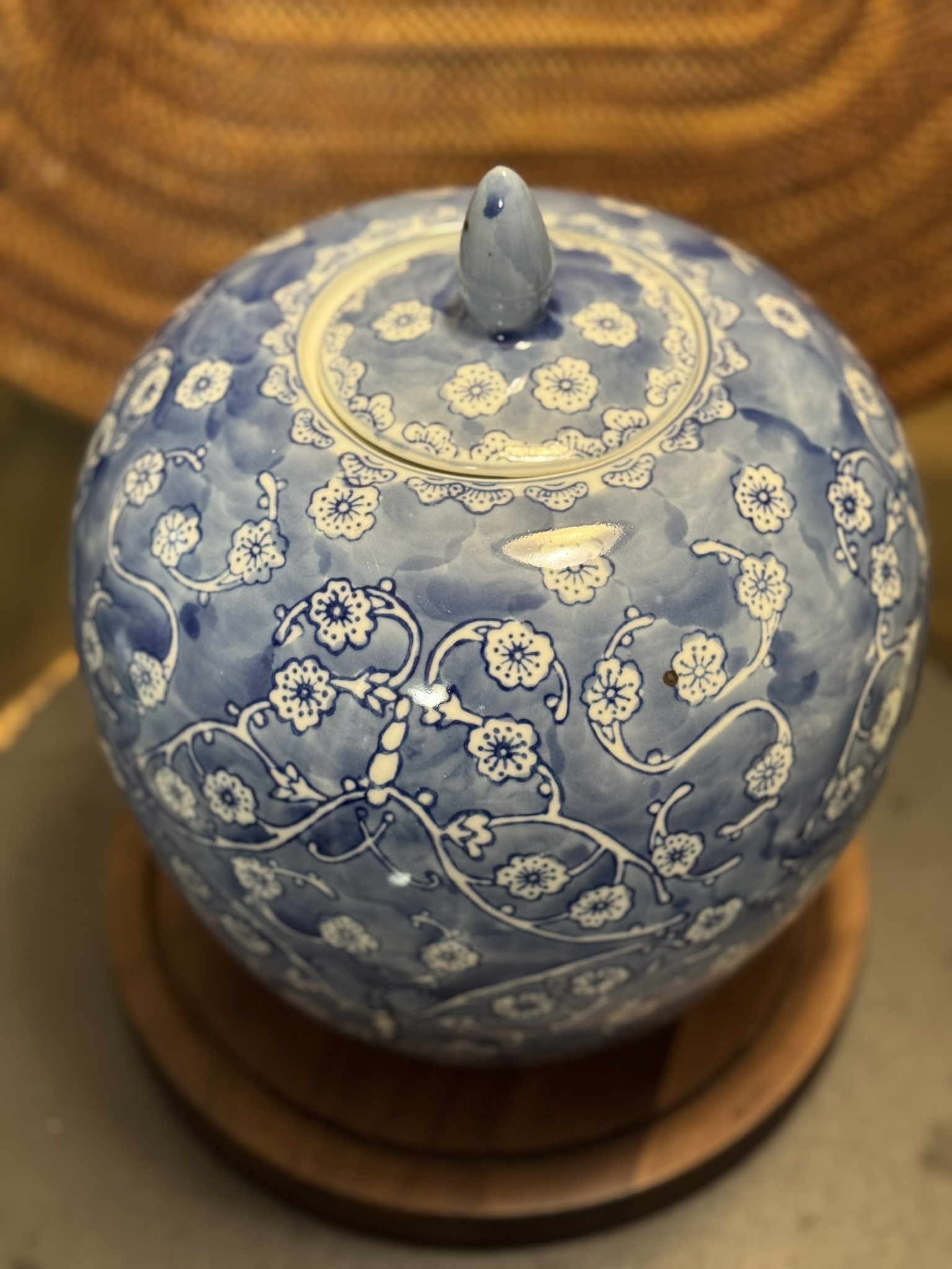 A round, blue ceramic jar with a lid sits on a wooden base. The jar is decorated with intricate white floral patterns and swirling vines. The lid has a small, pointed handle. The background is softly blurred in warm tones.