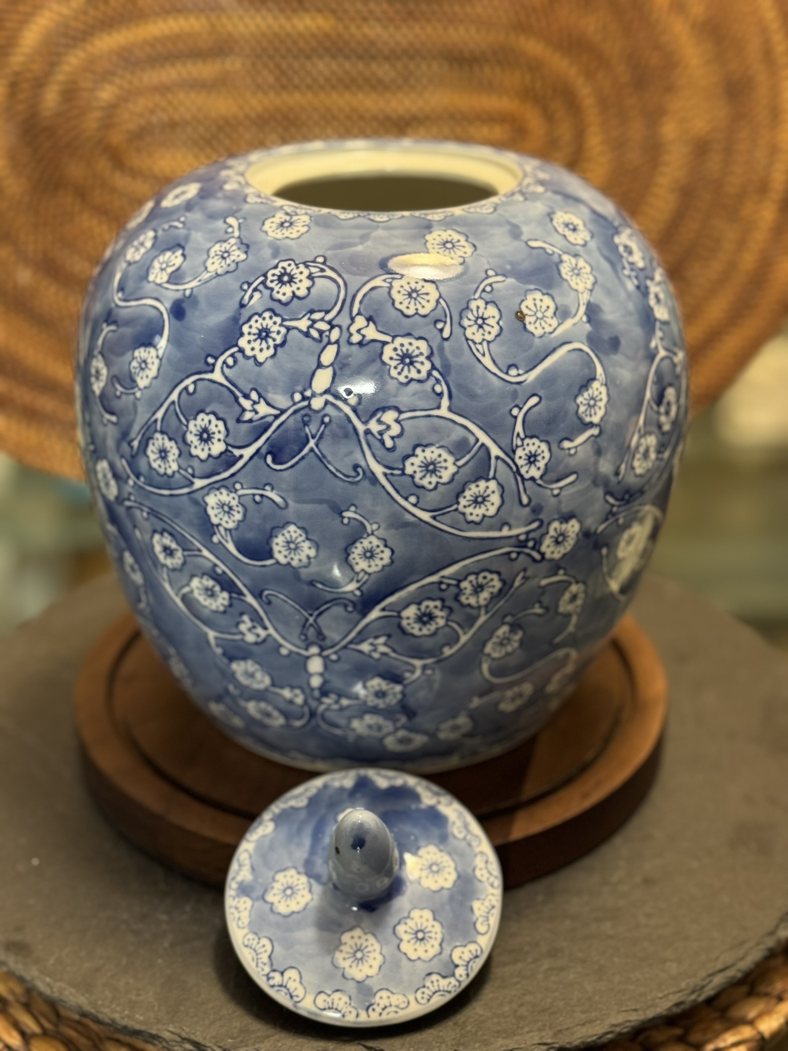 A blue and white ceramic jar with a floral pattern sits on a round wooden tray. Its lid, featuring the same pattern, is placed in front of the jar on the tray. The background shows a woven basket with a circular design.