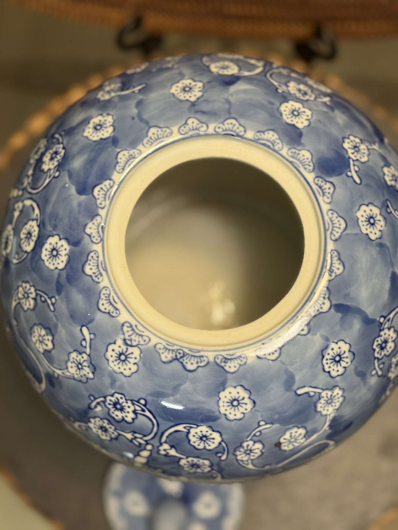 A close-up top view of a blue and white ceramic vase with a wide opening. The vase is decorated with delicate, repeating small white flowers and branches on a soft blue background. The photo is softly focused, and a coaster is partially visible underneath.