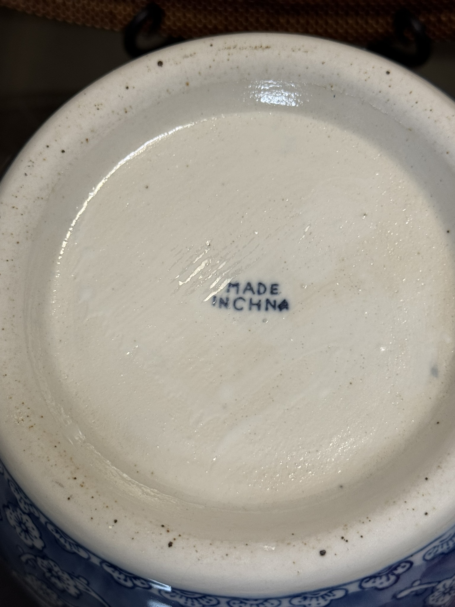 The bottom of a white ceramic object with blue floral patterns around the edge. In the center, MADE IN CHNA is stamped in blue, with CHINA missing the I. The surface is slightly textured with some brown specks.