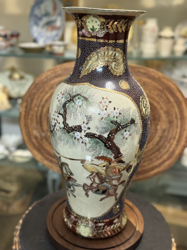A tall, ornate vase with a flared rim features a painted scene of warriors on horseback beneath a tree with blossoming flowers. The vase has intricate patterns in dark blue, gold, and cream, and is displayed in front of a woven circular mat on a shelf.