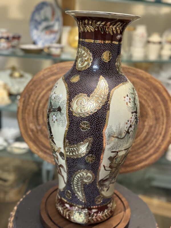 A tall, ornate ceramic vase features intricate paisley designs, gold leaf accents, and floral motifs. The background is dark with white dotted patterns. The vase is displayed on a wooden surface, with decorative plates and glassware blurred in the background.