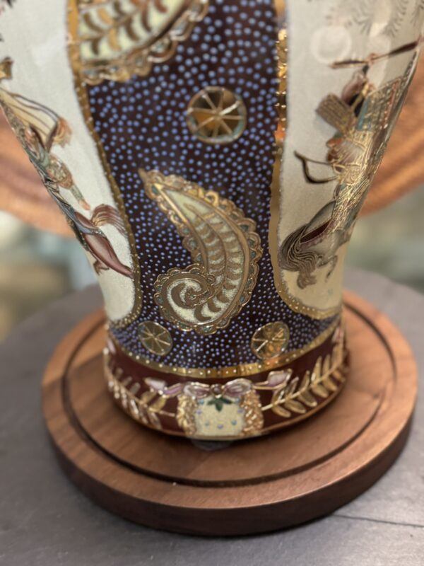 A close-up of a decorative vase with intricate gold designs, featuring paisley patterns, circular motifs, and detailed accents. The vase sits on a round wooden base, and some human figures are partially visible on its surface.