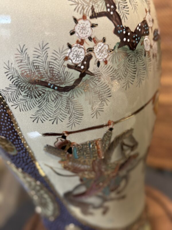 A close-up of a decorated ceramic vase shows a detailed painted scene: a samurai in ornate armor riding a horse beneath pine branches and blooming white flowers, all rendered with fine lines, muted earth tones, and metallic gold highlights.