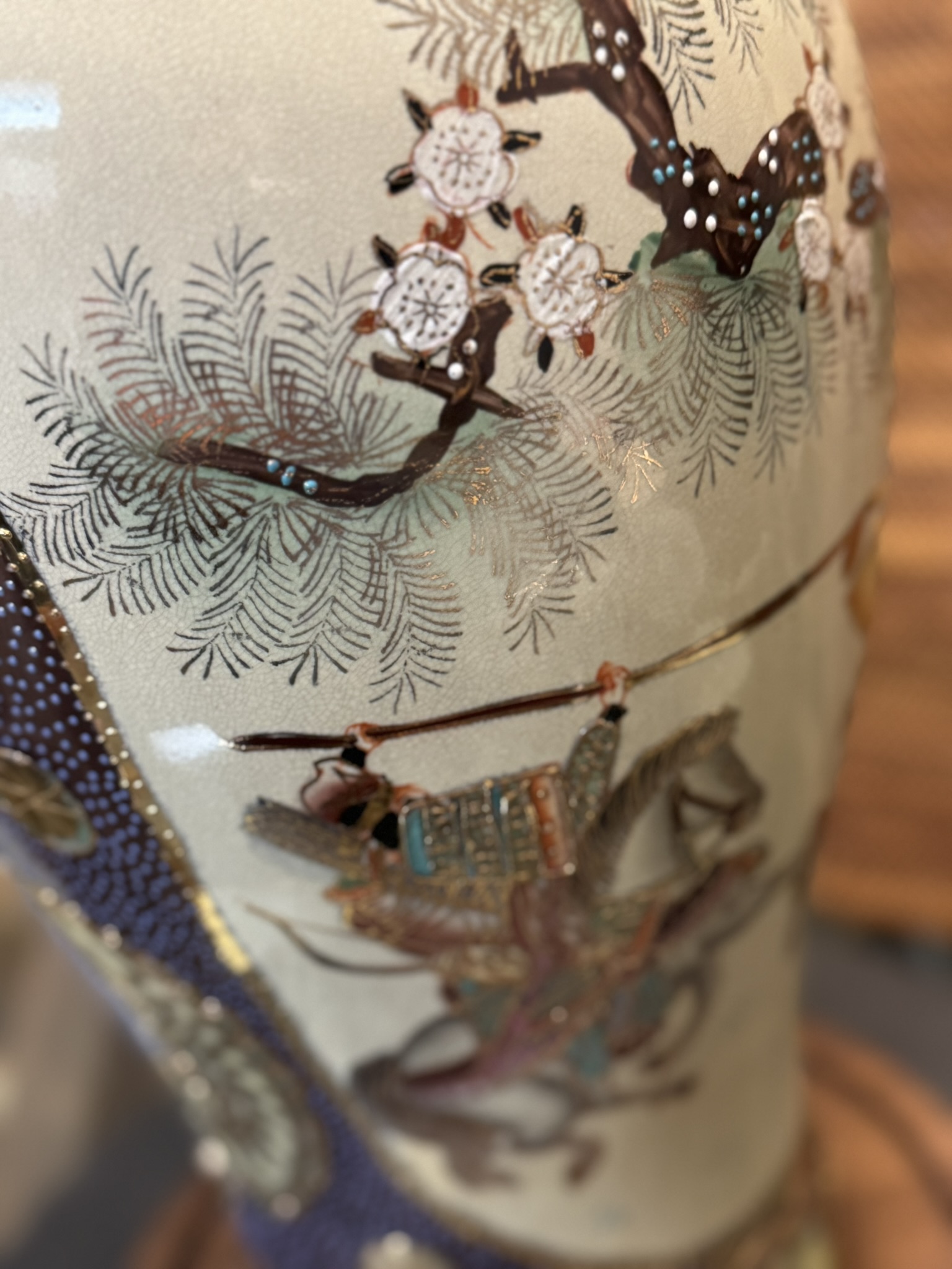 A close-up of a decorated ceramic vase shows a detailed painted scene: a samurai in ornate armor riding a horse beneath pine branches and blooming white flowers, all rendered with fine lines, muted earth tones, and metallic gold highlights.