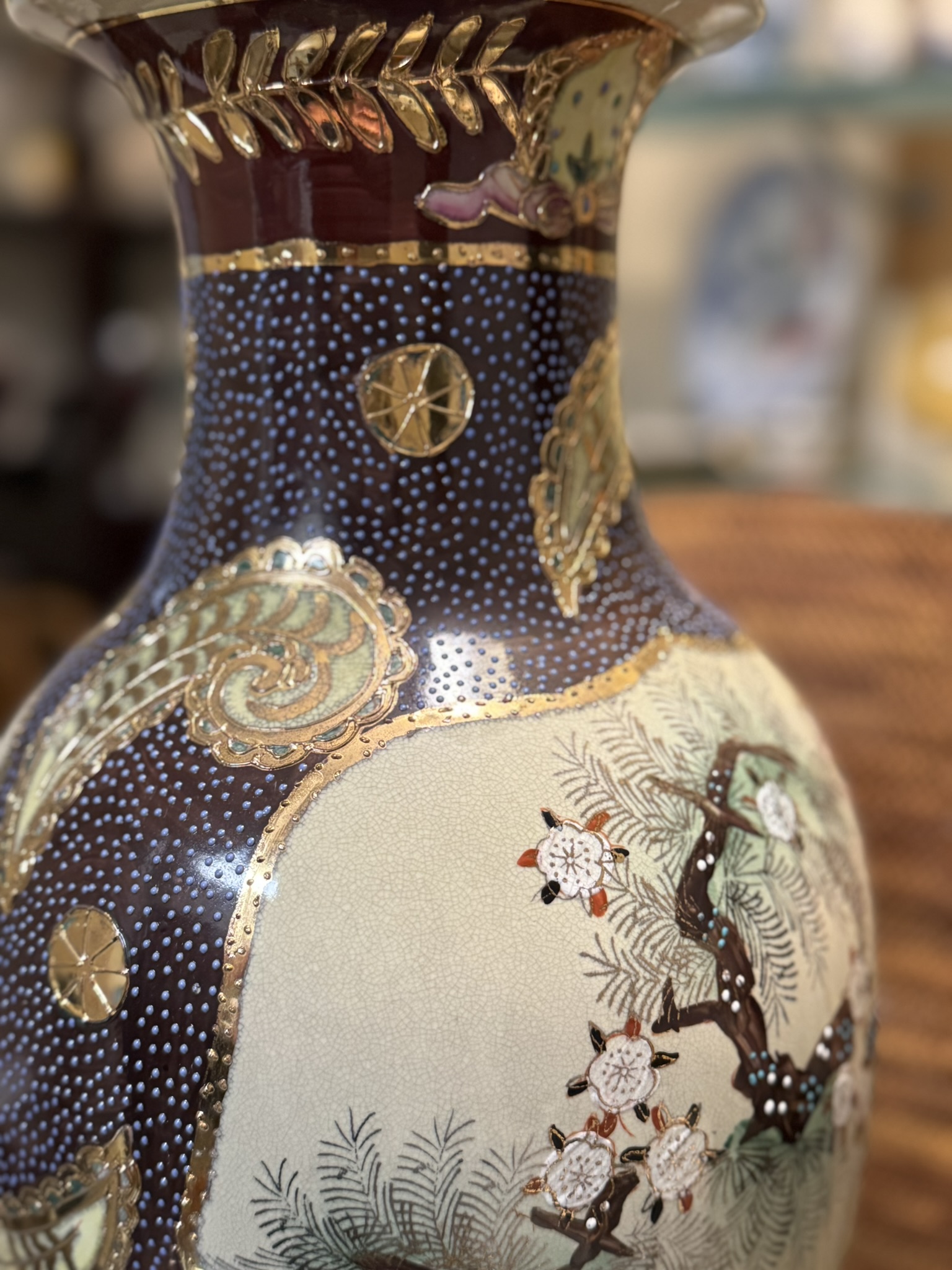 A close-up of an ornate ceramic vase featuring gold detailing, a maroon and blue patterned background, and painted pine branches with white blossom flowers. The vase has intricate textures and decorative motifs highlighted with metallic accents.