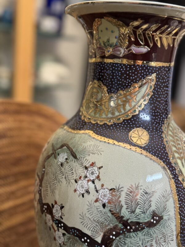 A close-up of a decorative vase with intricate patterns. The vase features a painted branch with white blossoms and textured designs in gold, maroon, and green. The background shows a wicker chair and a blurred shelving unit.
