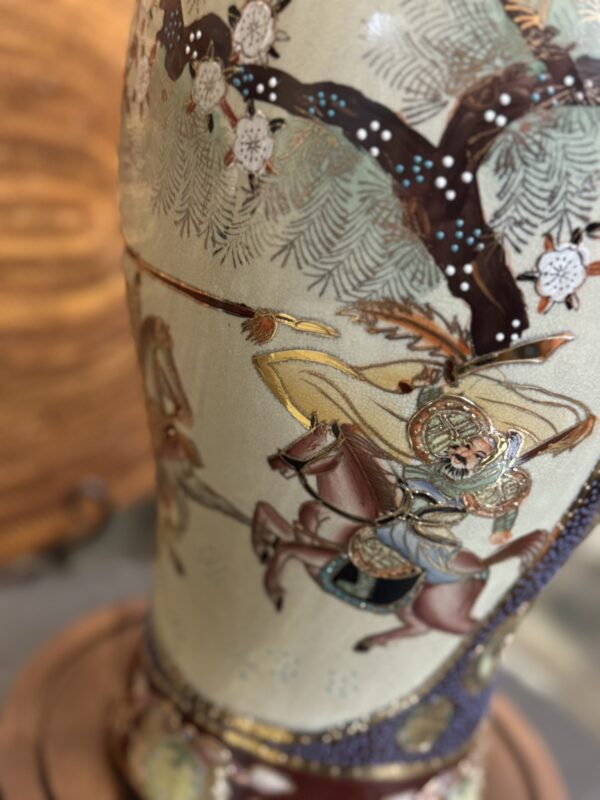 A close-up of a ceramic vase decorated with an intricate scene of an armored warrior on horseback, holding a spear under a blooming tree with white flowers. The background is blurred, highlighting the vase’s detailed artwork.