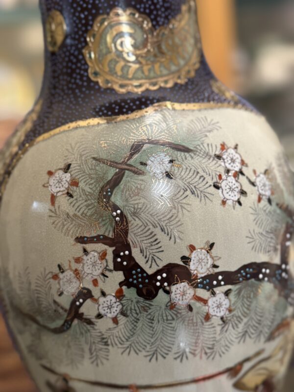 A close-up of a decorative ceramic vase featuring a painted design of a dark, twisting tree branch with white blossoms and delicate green leaves on a cream background. The upper part of the vase is deep blue with gold accents.