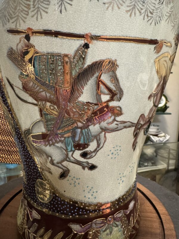 A close-up of a decorative vase features a detailed painting of an armored horseman wielding a spear, riding a galloping white horse. The vase displays rich colors, gold accents, and intricate patterns, with another figure partially visible on the right.