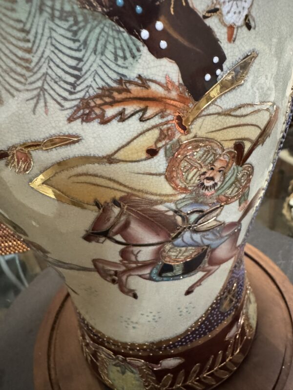 A detailed close-up of a ceramic vase depicts a warrior in ornate armor riding a horse. The scene includes intricate patterns, gold accents, swirling lines, and foliage, all painted in muted tones atop a textured, crackled surface. The vase sits on a wooden base.