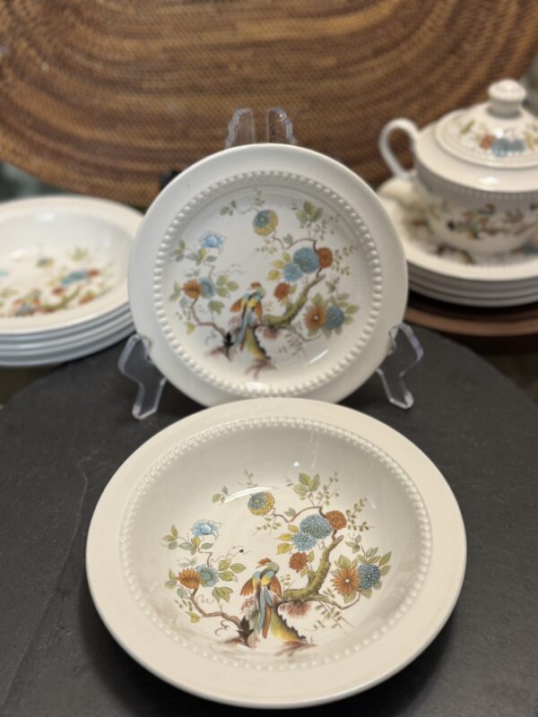 Two white ceramic plates with a raised dotted border and colorful bird and floral designs are displayed upright. In the background, stacked matching plates and a teapot with a lid are visible, all set against a woven textured backdrop.